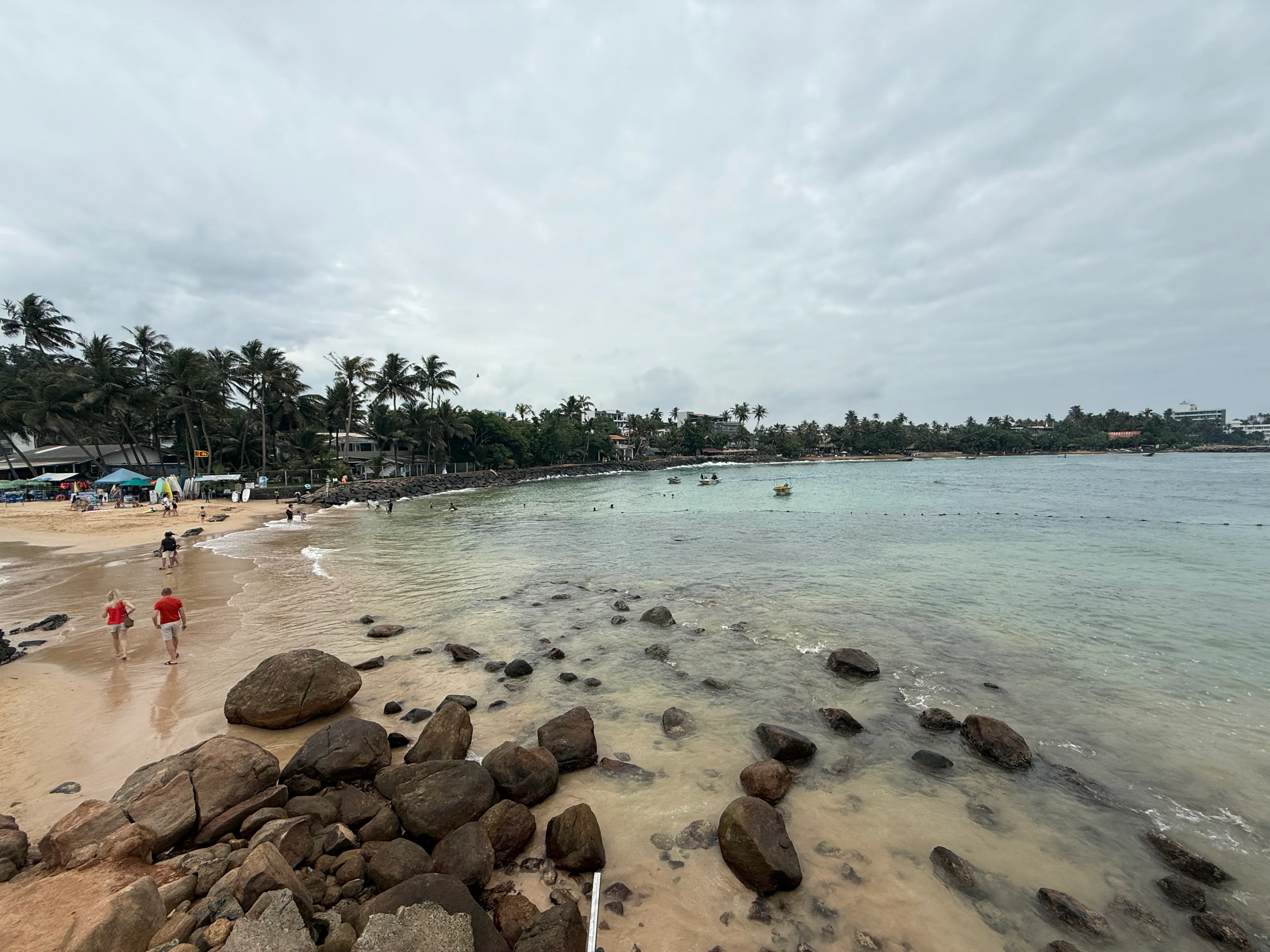 Mirissa Bay beach and calm sea under low monsoon cloud, palms and shore rocks along the sand