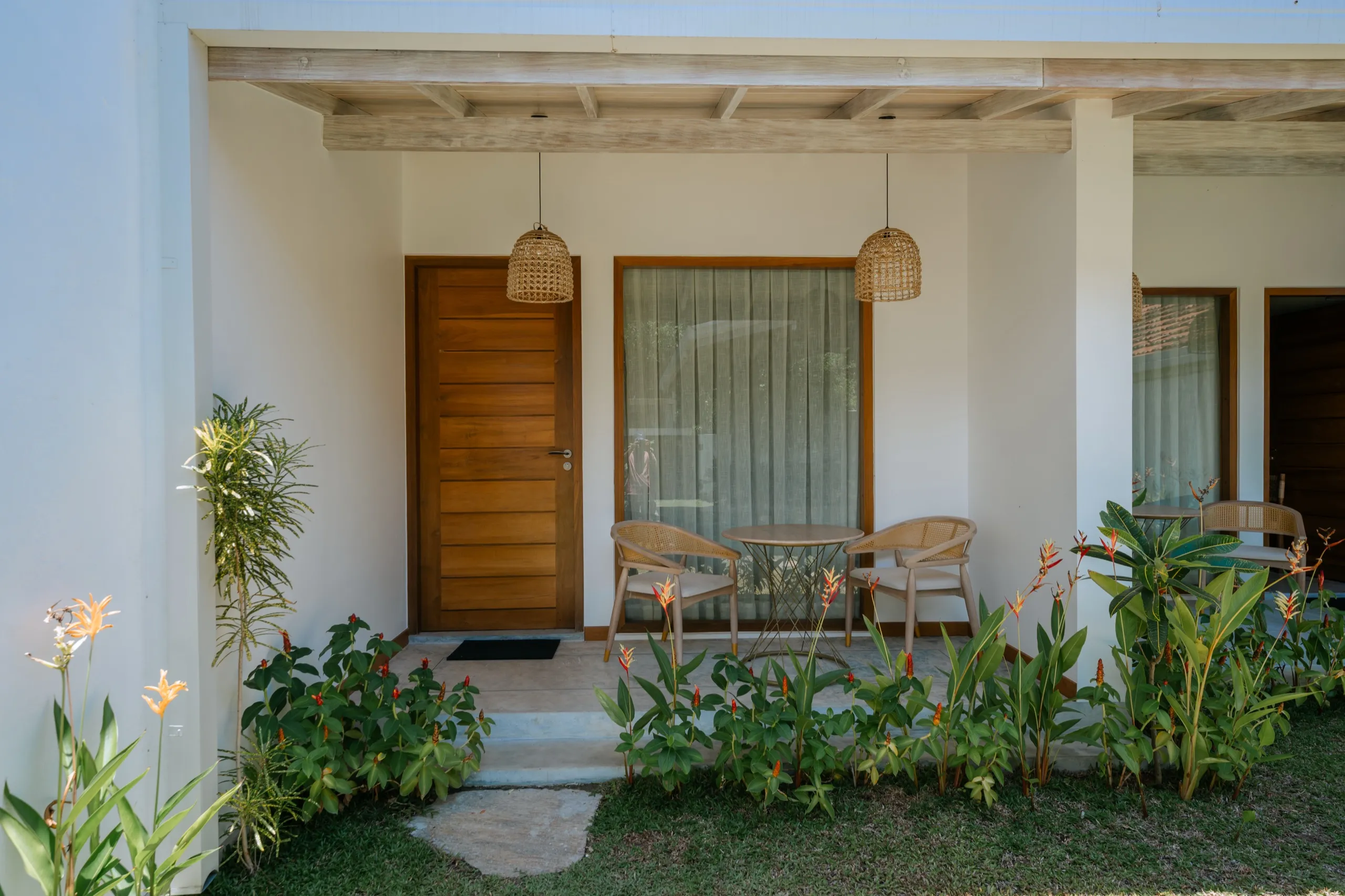 The entrance to a new villa room: timber door and curtained glass wall, two cane chairs and a small table on the terrace under woven lanterns, tropical planting in front