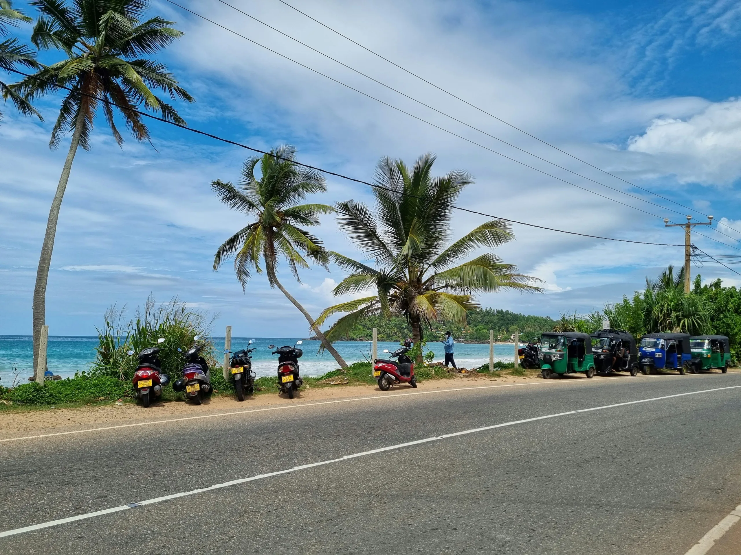 Scooters and tuk-tuks parked under coconut palms along the coastal road at Mirissa, the ocean visible behind