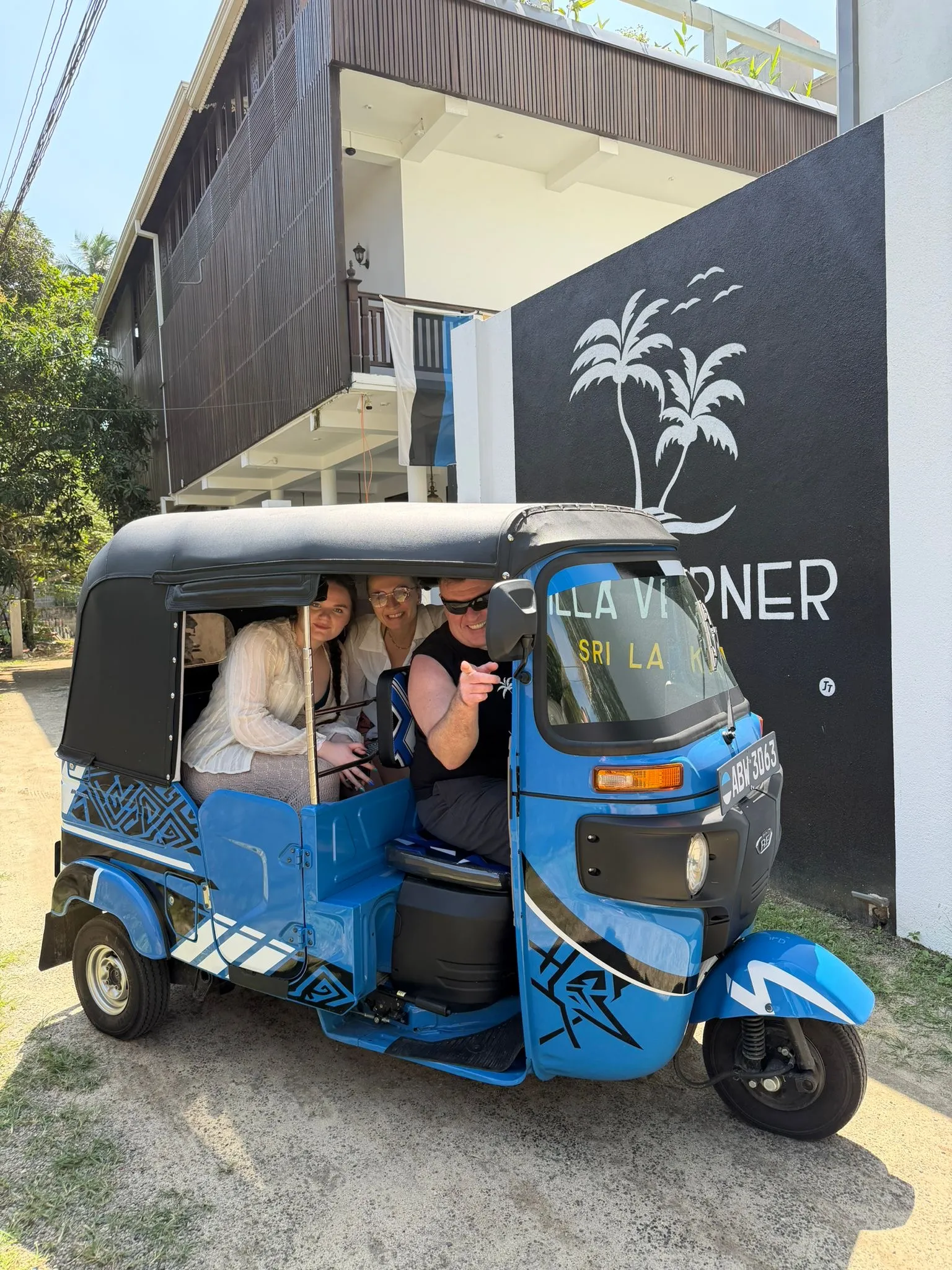 A blue tuk-tuk on the villa drive, the Villa Verner palm mural painted on the dark wall behind it